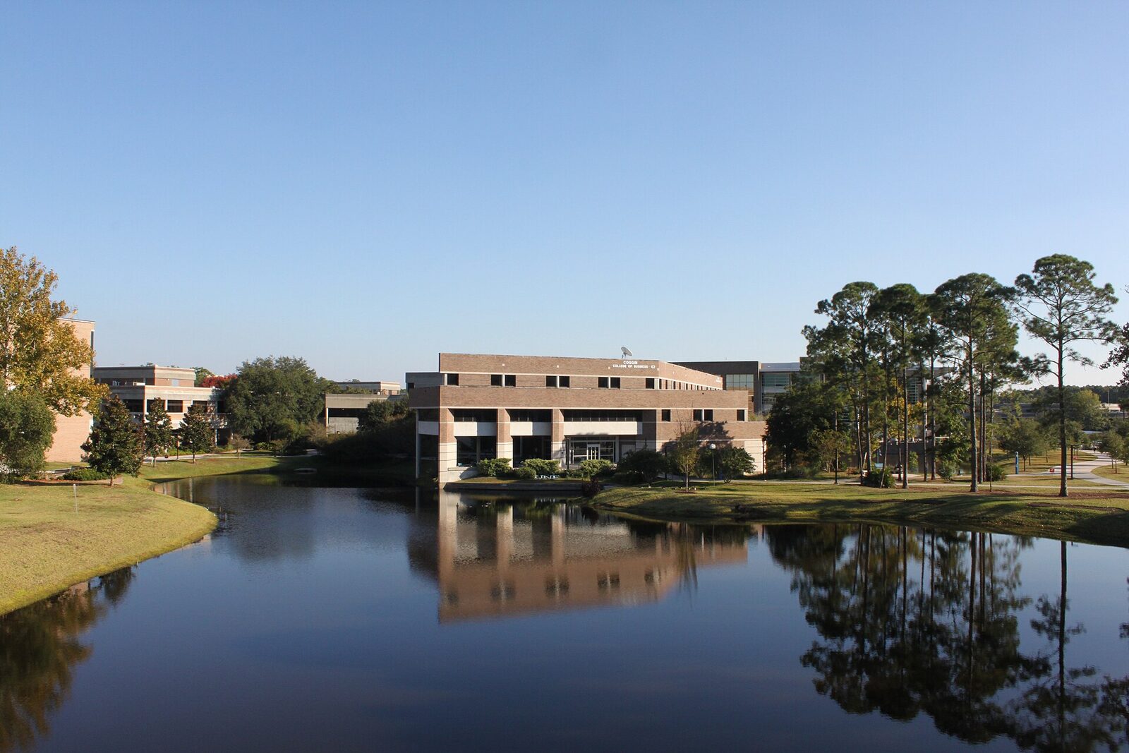 A low brick university building and pine trees reflected in a still lake on the University of North Florida campus
